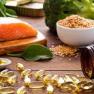 alt="A close-up of nutrient-rich foods and supplements on a wooden surface, featuring a raw salmon fillet on a cutting board, a bowl of sesame seeds, fresh spinach leaves, broccoli, and a white spoon containing olive oil and an olive. Nearby, an amber bottle is tipped over, spilling golden omega-3 capsules."