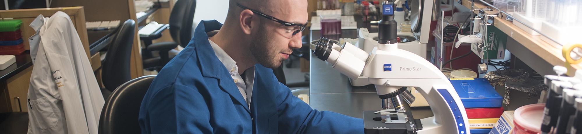 Male student in PPE in lab setting using a microscope
